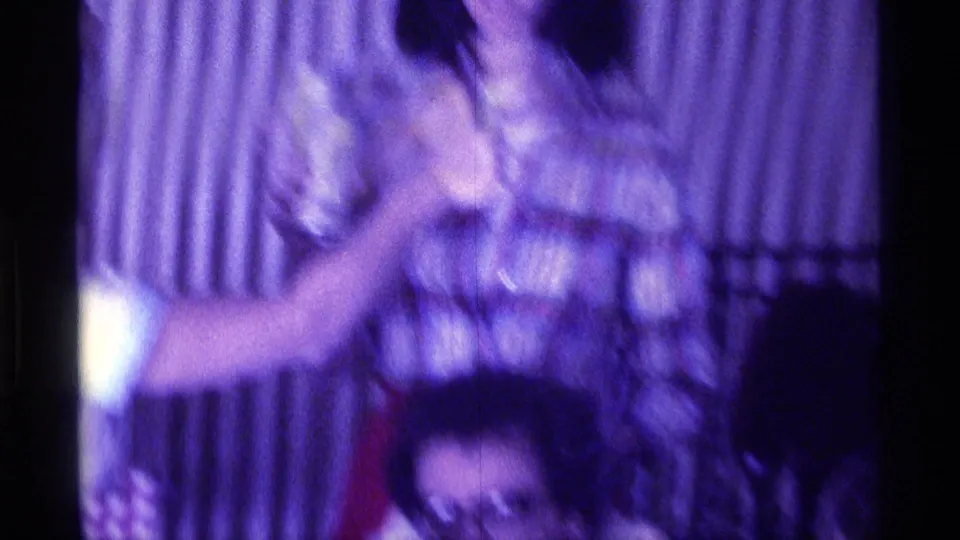 Blurred 1976 Kentucky home-movie frame of an adult with a microphone moving in front of curtains during a family sing-along moment.