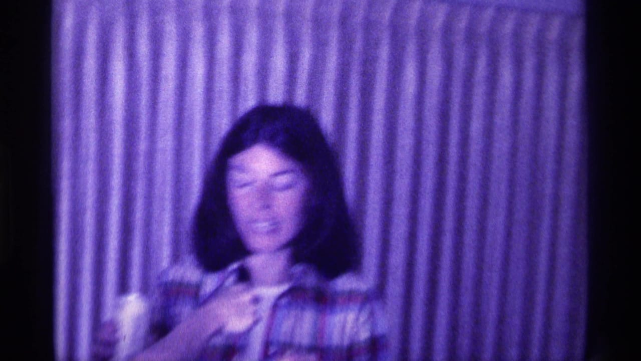 Young woman in a plaid shirt holds a drink in front of striped curtains.