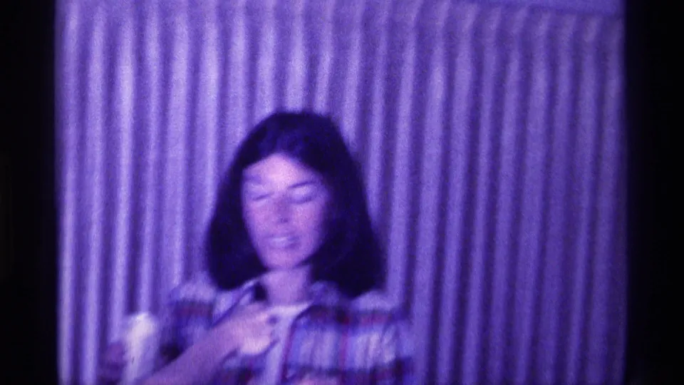 In 1976 Kentucky home-movie footage, a young woman in a plaid shirt appears to speak while holding a drink before striped curtains.