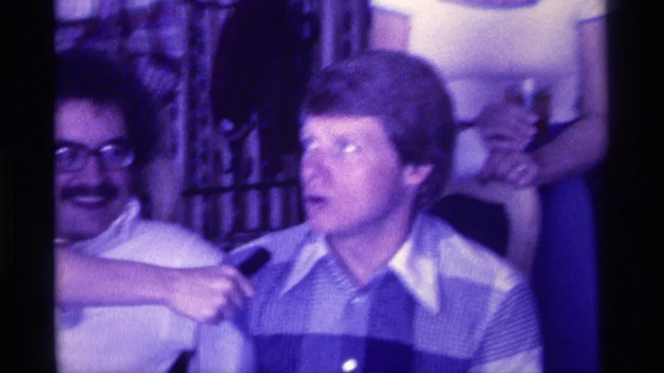 A younger man in a checkered shirt appears to answer a handheld microphone while another man in glasses sits nearby in the private room.