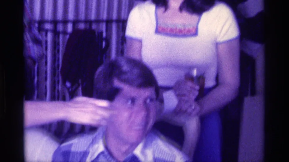 A seated man looks upward as a hand extends a microphone toward him, with other adults partly visible behind him in the room.