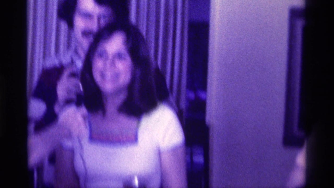 Man with mustache behind smiling woman in white top inside a curtained room.