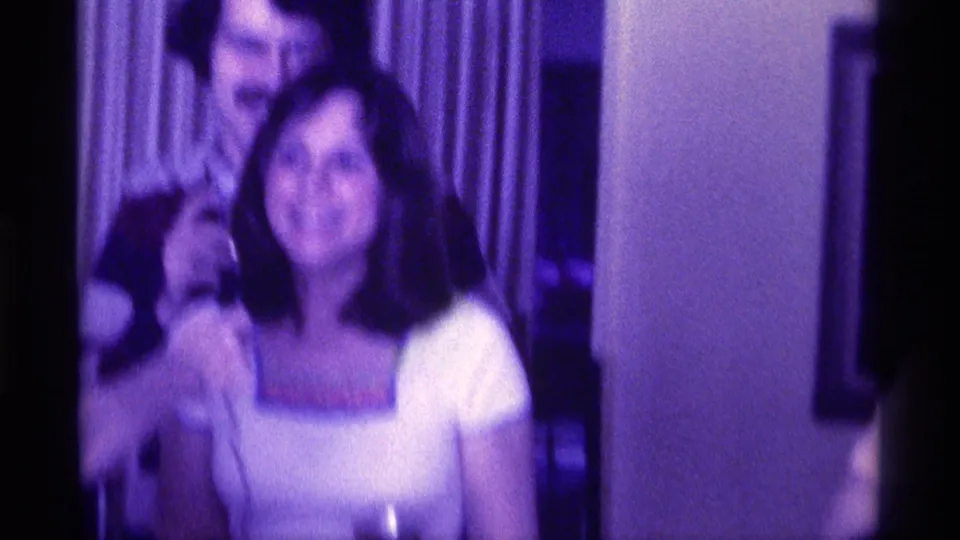 1976 Kentucky home-movie frame showing a mustached man standing behind a smiling woman in a white top during an indoor gathering.