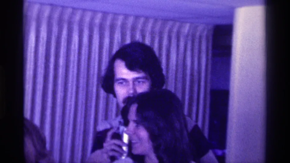 1976 Kentucky home-movie frame of a mustached man holding a drink beside a woman during an indoor social gathering.