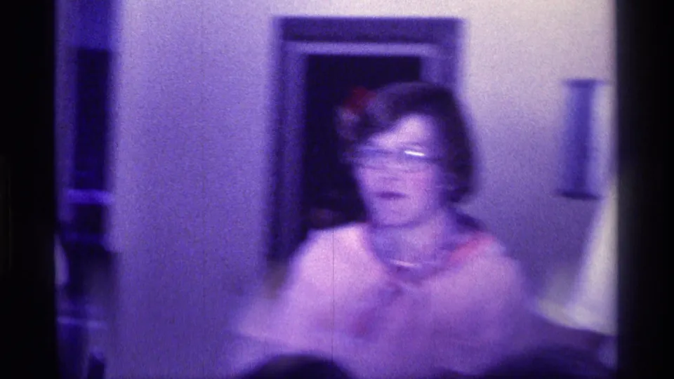 1976 Kentucky home-movie frame showing a woman with glasses and a scarf in soft motion blur inside a private room.