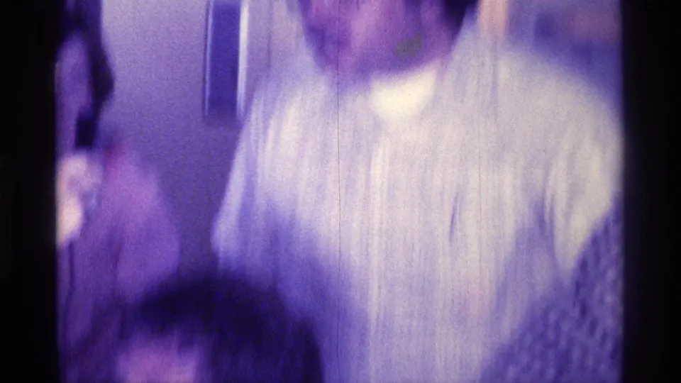 1976 Kentucky home-movie frame of a woman in a light patterned top moving through an indoor gathering with other adults nearby.