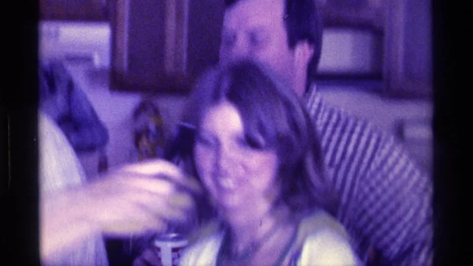 1976 Kentucky home-movie frame showing adults at an indoor party, with a woman holding a cup and a man in a striped shirt nearby.