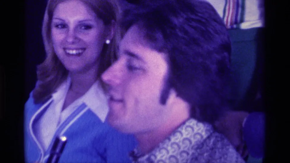 1976 Kentucky home-movie frame showing a smiling woman behind a man in a patterned shirt with a handheld microphone.