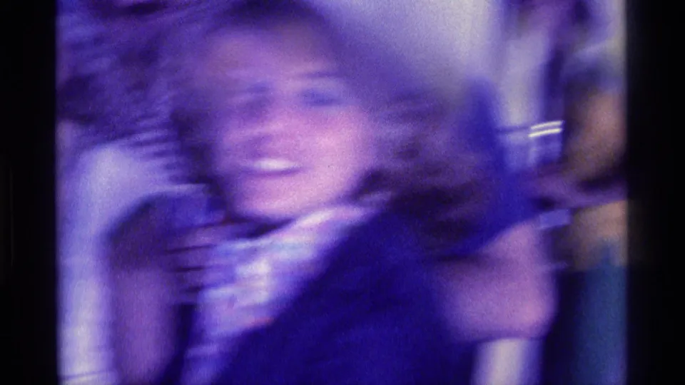 Heavily blurred 1976 Kentucky home-movie frame of an adult at an indoor party, with a purple cast and indistinct drink gesture.