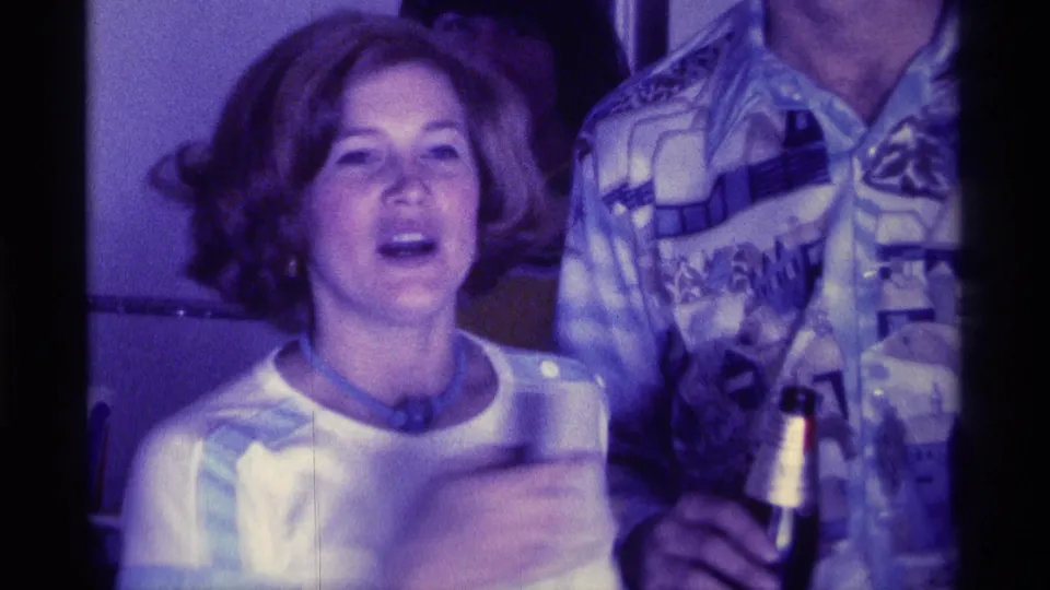 1976 Kentucky 8mm party frame showing a woman in a light top holding a cup beside a man in a patterned shirt inside a private room.