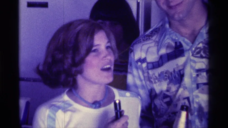 1976 Kentucky home-movie frame of a woman holding a drink and looking toward a man in a patterned shirt during an indoor party.