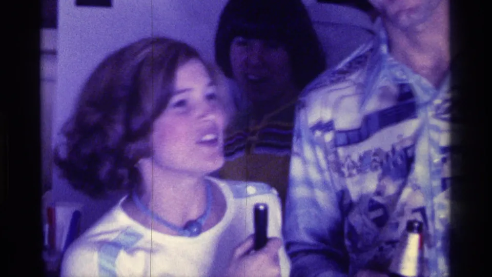 The woman appears mid-sentence or reacting with a microphone in hand beside a patterned-shirt man in a Kentucky home-movie interior.