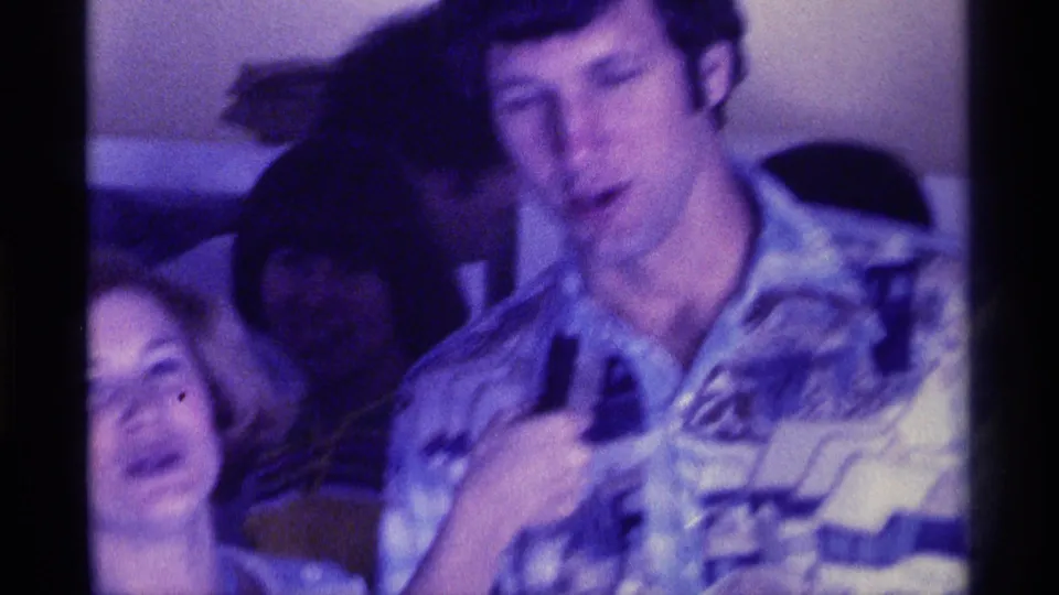 A man in a patterned shirt holds the microphone during the indoor 1976 Kentucky gathering, with nearby adults softened by motion blur.