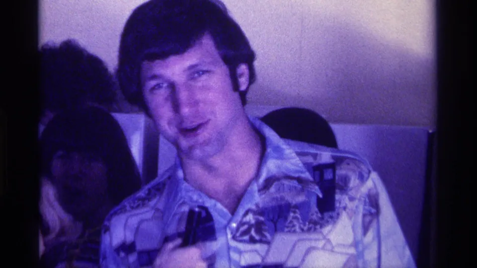 A smiling man with a microphone faces the camera in a dim private interior, with other adults partly visible behind him.