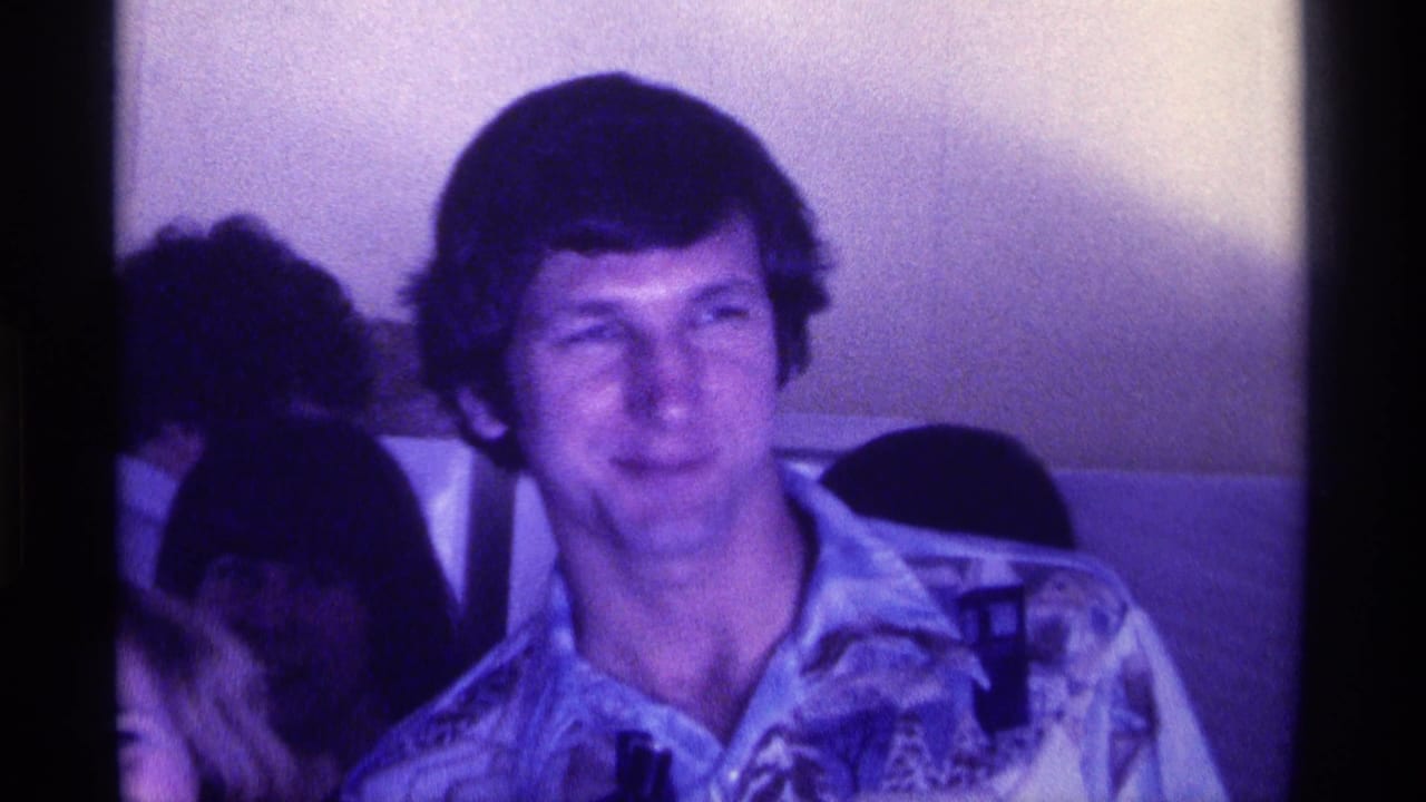 In 1976 Kentucky home-movie footage, a young adult man faces the camera in a crowded dim interior with other people close by.