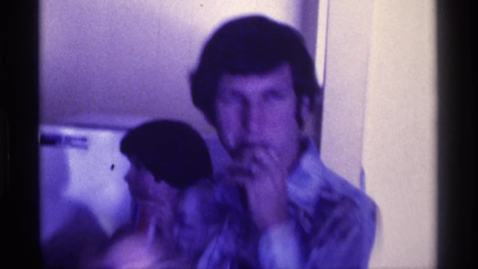 In 1976 Kentucky footage, a mustachioed adult man holds a microphone near his mouth while people stand behind him indoors.