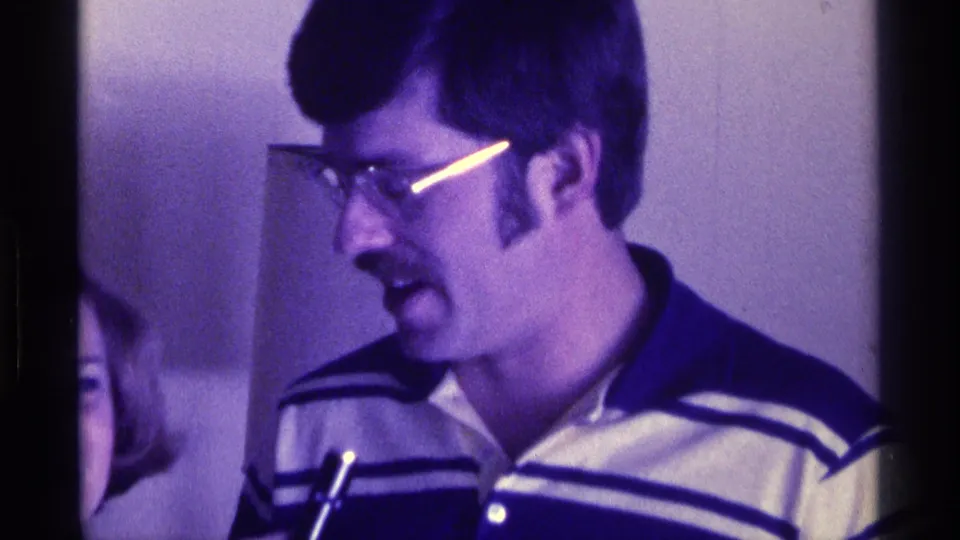 A mustachioed man in glasses and a striped polo shirt appears in profile beside a microphone in 1976 Kentucky home-movie footage.