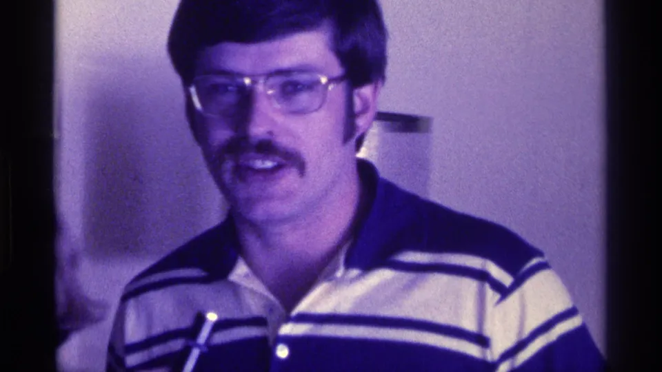 A mustachioed man wearing glasses and a striped polo faces the camera as a microphone is visible below in a 1976 Kentucky interior.