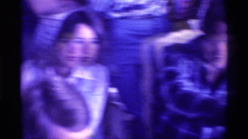 1976 Kentucky home-movie frame with a seated woman facing forward amid a crowded family viewing scene.