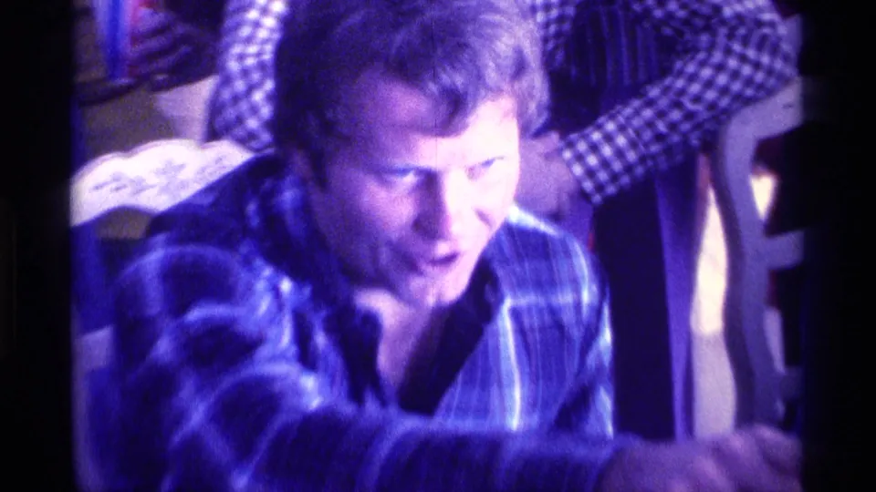 1976 Kentucky 8mm frame showing a man in plaid leaning forward with visible anticipation in a private family room.