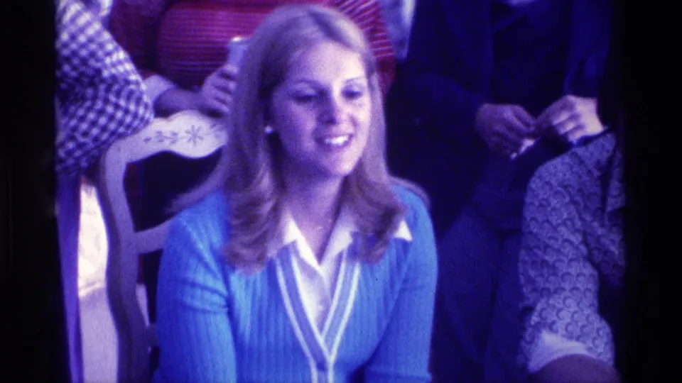 1976 Kentucky home-movie frame of a smiling woman in a blue cardigan seated with others during a family viewing moment.