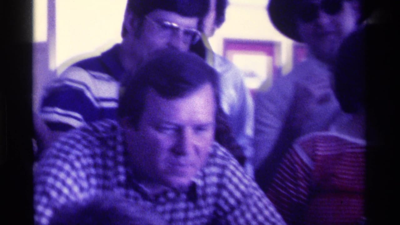 Adults in a Kentucky 1976 home-movie frame lean together indoors while watching a horse race on television.