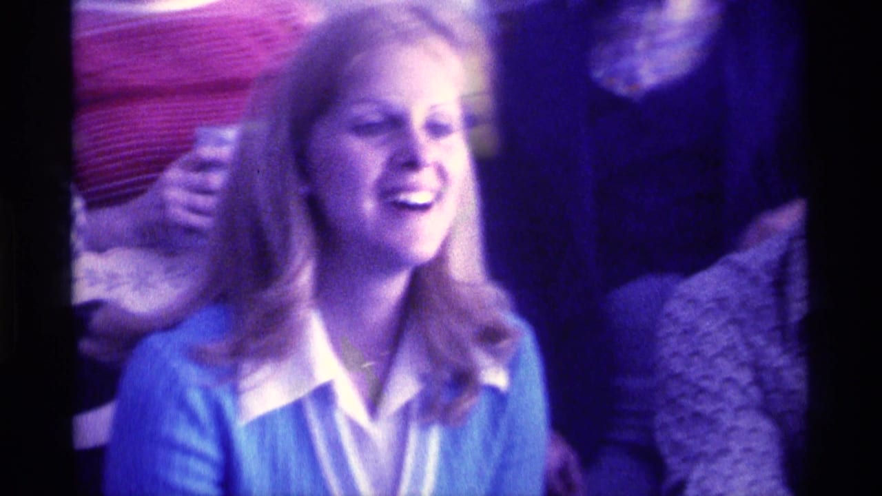 1976 Kentucky 8mm frame of a blonde woman in a blue cardigan laughing near other adults in a crowded indoor gathering.