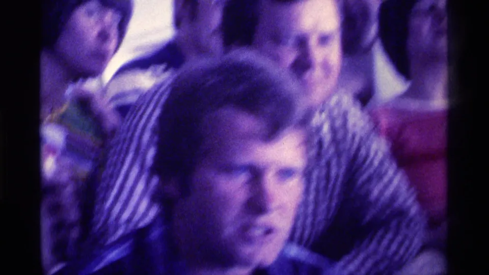 1976 Kentucky 8mm frame with a young man in the foreground and several seated adults behind him in a crowded private interior.