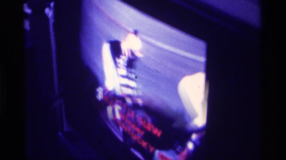 Television viewed in a dark room shows blurry horse-racing footage with red broadcast text and strong purple-blue color shift.