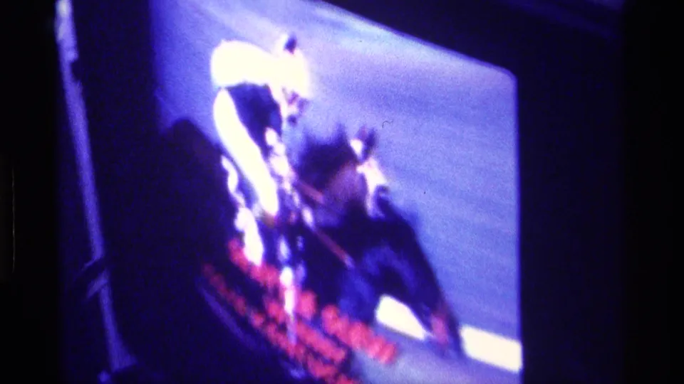 Close home-movie view of a television race broadcast, with a jockey and horse shape visible through blur and red on-screen text.