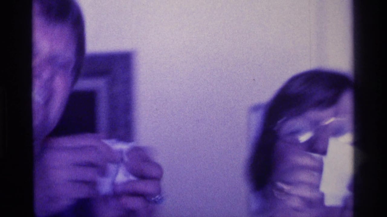Two adults in a dim room, one holding a small object and one wearing glasses.