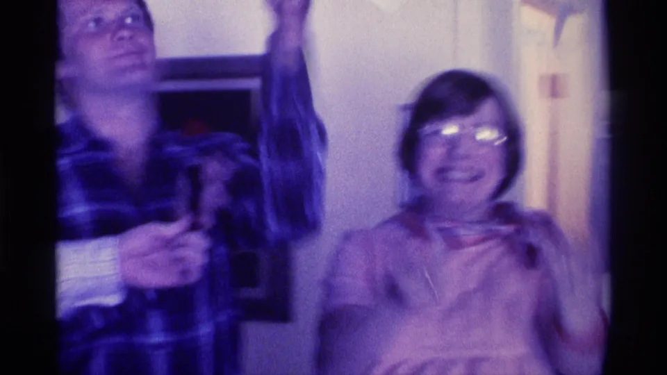 Kentucky 1976 8mm frame of two adults in a private room, with one person raising an arm and another smiling in glasses.
