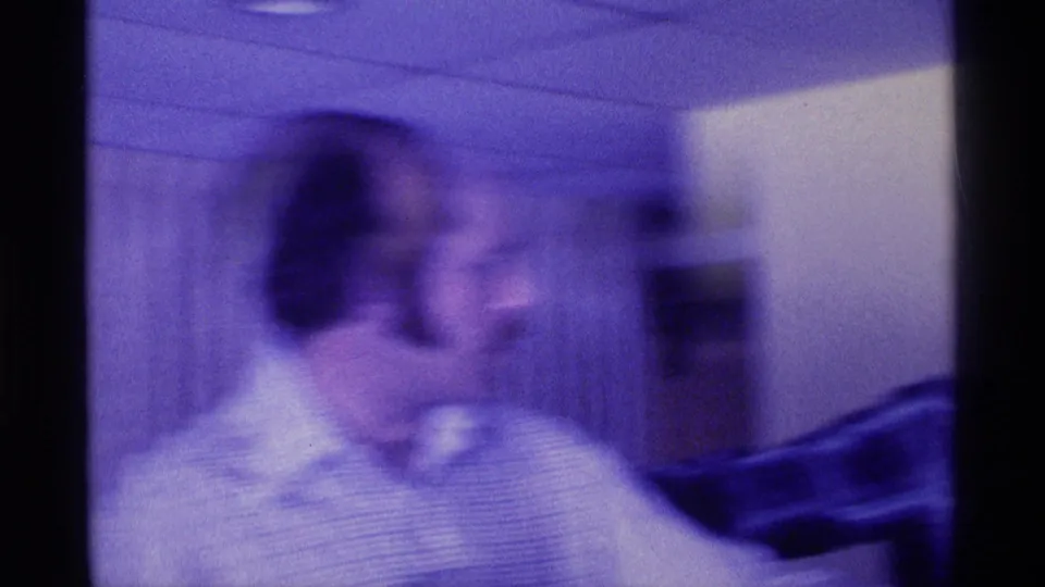 Kentucky 1976 home-movie frame showing a blurred adult man in profile in a private interior with dark furniture nearby.