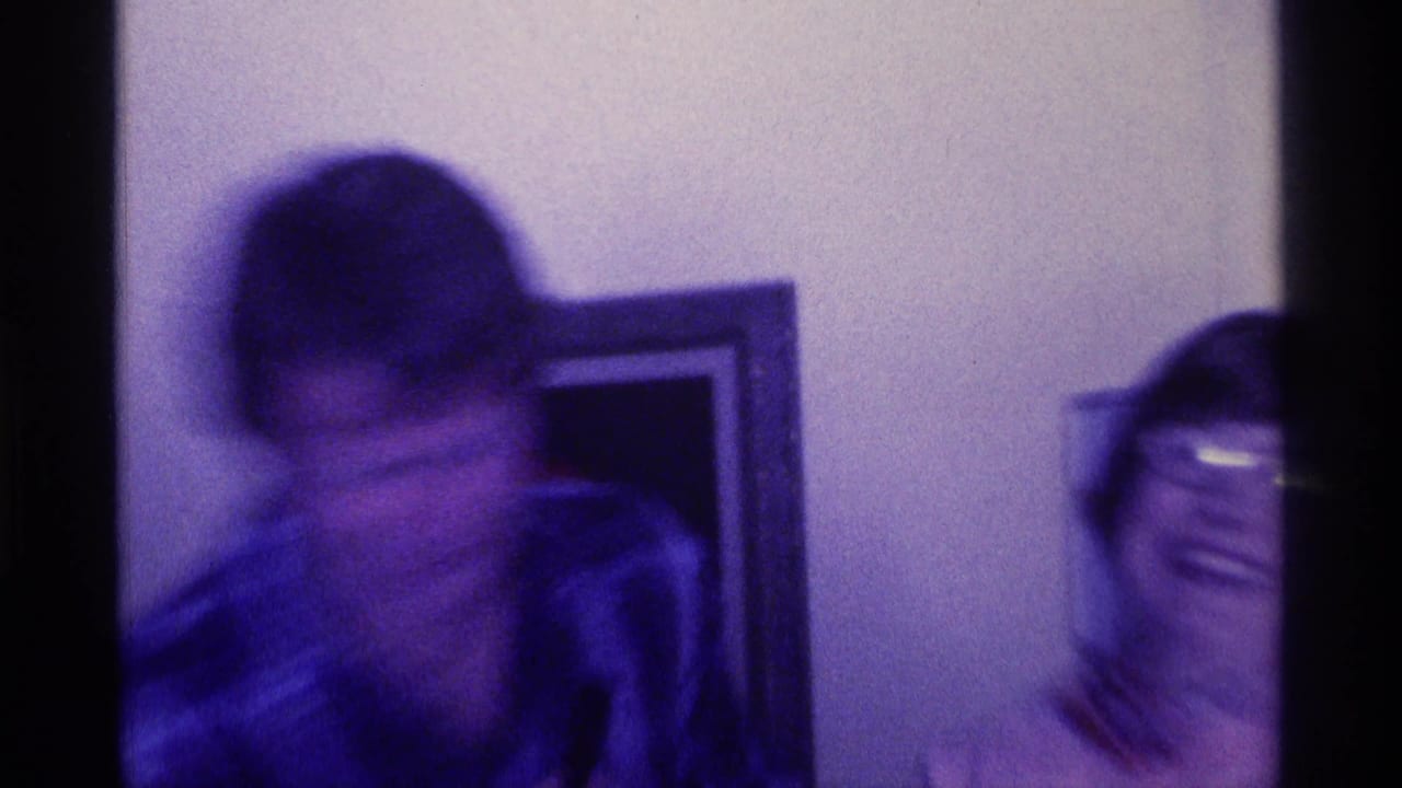 Motion-blurred 1976 Kentucky home-movie frame with a man moving near a laughing woman in glasses inside a gathering space.