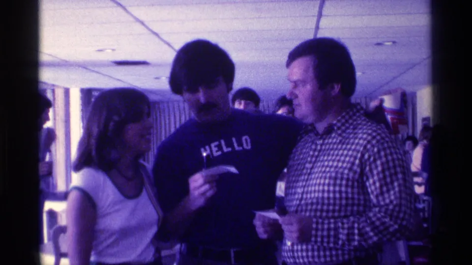 1976 Kentucky 8mm frame of adults talking indoors, including a man in a HELLO shirt and another in a checkered shirt.