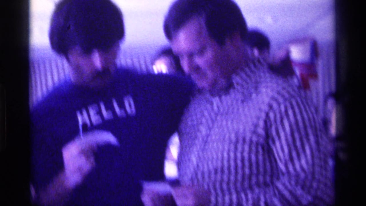 A man in a dark HELLO shirt turns toward a smiling man in a checkered shirt inside a private-looking 1976 Kentucky interior.