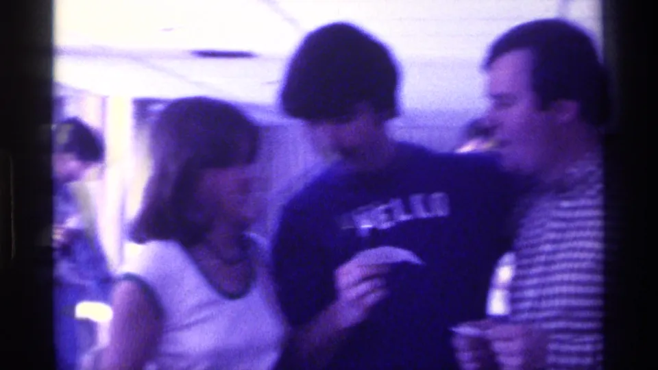 The woman in a white top faces the mustached man in the HELLO shirt, with another man smiling nearby in the 1976 Kentucky home-movie scene.
