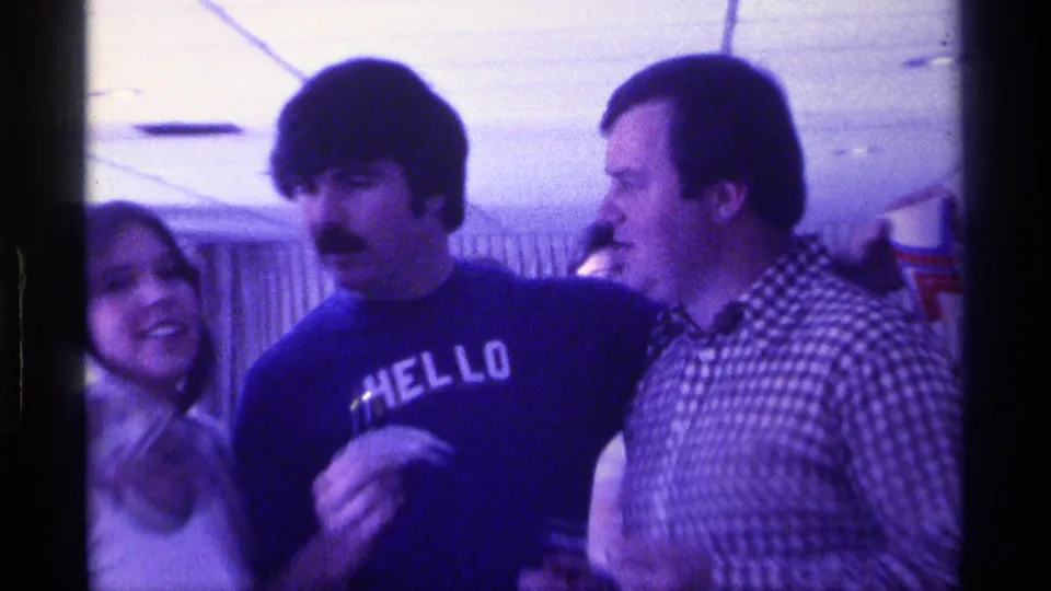 Three adults smile in a 1976 Kentucky interior as the center man in a HELLO shirt holds small paper bills or slips near a man in a checkered shirt.