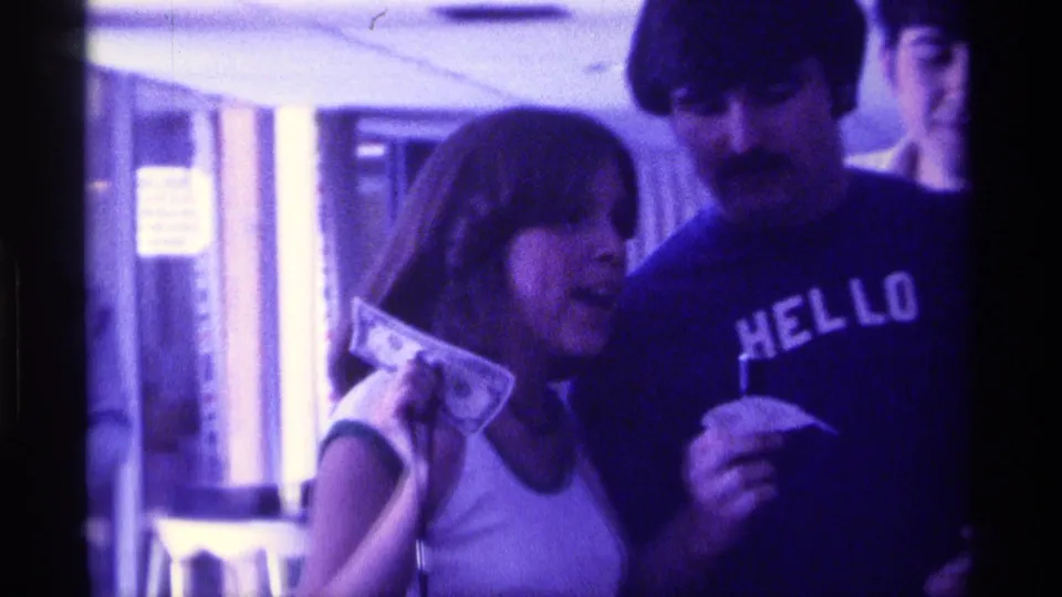 A woman holds paper currency near the man in the HELLO t-shirt, with curtains and other adults visible in the 1976 Kentucky interior.