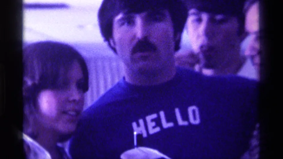 The HELLO t-shirt is clearest as the man faces the camera, with a woman and another man crowded close in the indoor gathering.