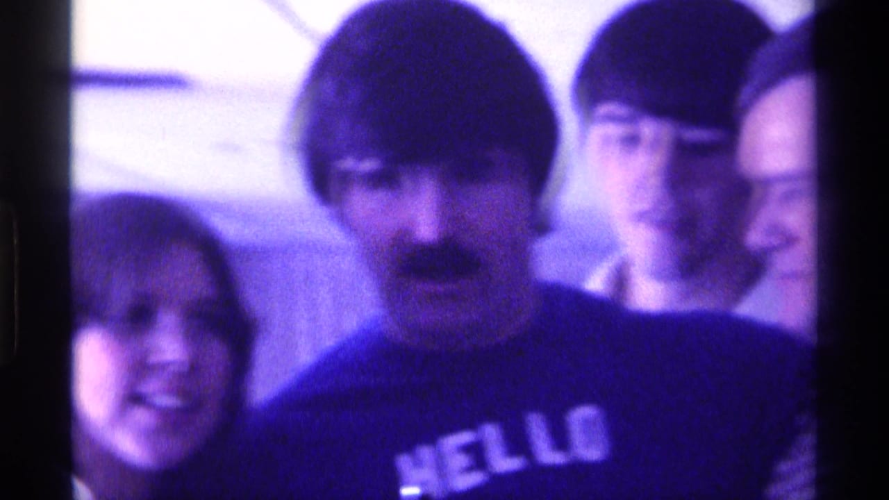 1976 Kentucky home-movie frame of adults at an indoor party, centered on a mustached man in a dark HELLO shirt.