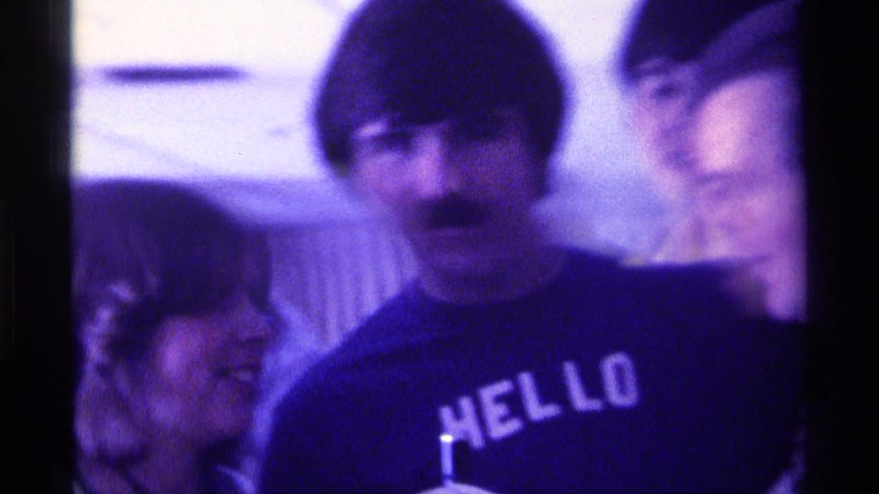 Woman smiles beside a mustached man in a dark HELLO shirt as others stand nearby.