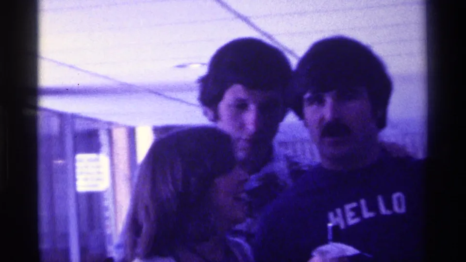 1976 Kentucky party frame showing three adults in close conversation, with curtains and ceiling panels visible behind them.