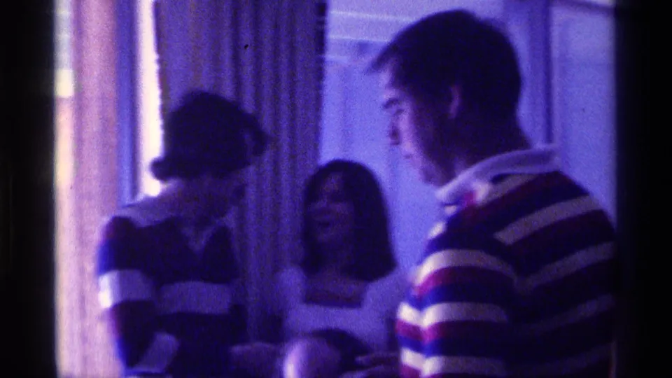 Indoor 1976 Kentucky home-movie frame of party guests talking near curtains, including two striped sweaters and a bright top.