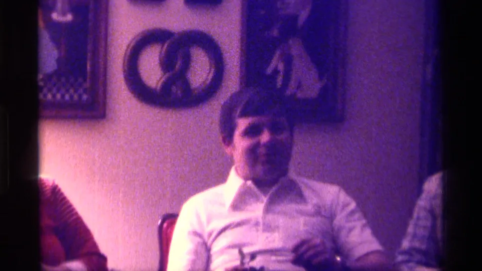 A man in a white collared shirt sits in a private Kentucky interior with framed wall art and a pretzel-shaped wall decoration behind him.
