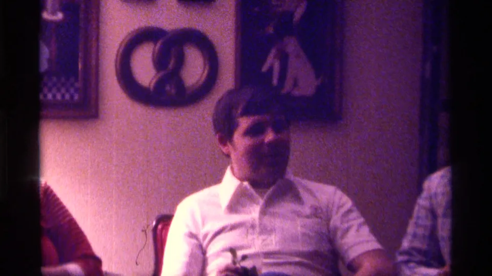 A seated man in a white collared shirt is shown in the same private interior, with another person partly visible at the right edge.