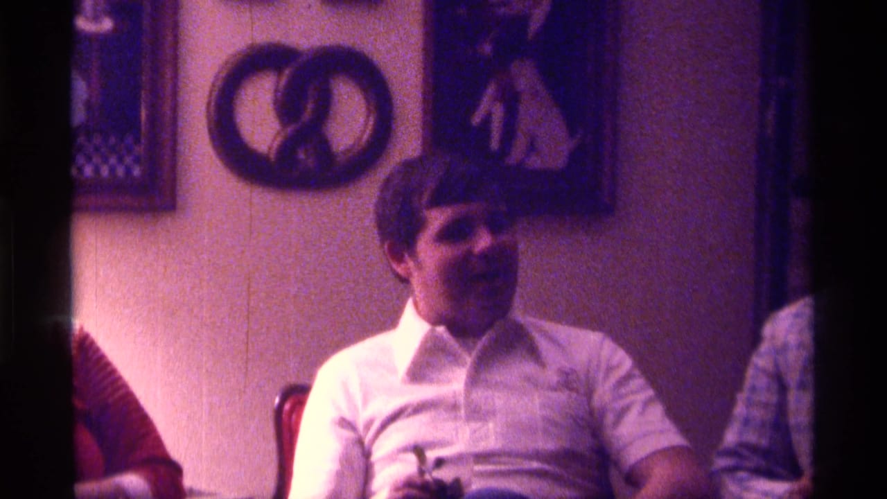 Adult man in a white collared shirt sits in a dim room with wall art behind him.