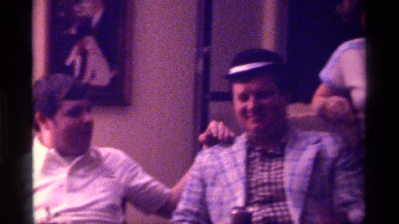 Two adult men sit together in a 1976 Kentucky interior, one in a white shirt resting his hand on a man wearing a plaid jacket and bowler-style hat.