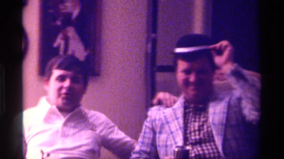 The man in the plaid jacket lifts his hat brim toward the camera while seated beside a man in a white shirt in the 1976 Kentucky home-movie scene.