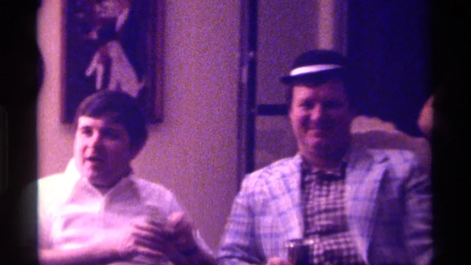 Two adult men face the camera in a dim 1976 Kentucky interior, with the man in the plaid jacket and hat holding a drink can after the hat-tip gesture.